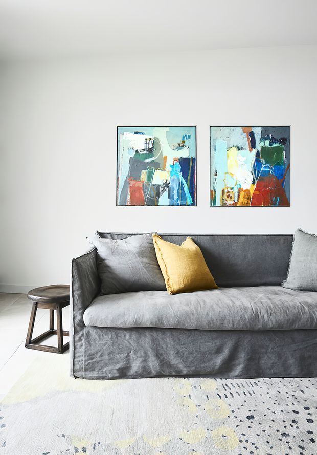 A bright, minimalist living room featuring a large grey slipcovered sofa, a small dark timber side table, a pale patterned area rug, and two colourful abstract artworks mounted on a white wall. The space has a calm, contemporary feel with yellow and blue accents.
