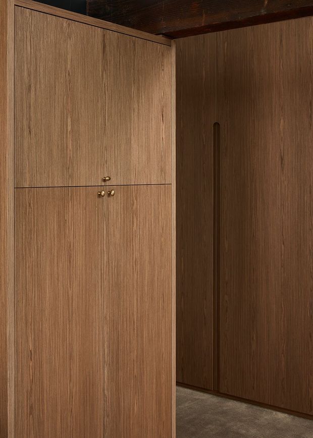 Close-up interior view of floor-to-ceiling dark timber built-in cabinetry and wardrobe doors, with vertical wood grain, integrated recessed handles, and small brass knobs. The cabinetry wraps around a corner beneath a dark timber ceiling, with a muted grey-brown floor and moody warm lighting.