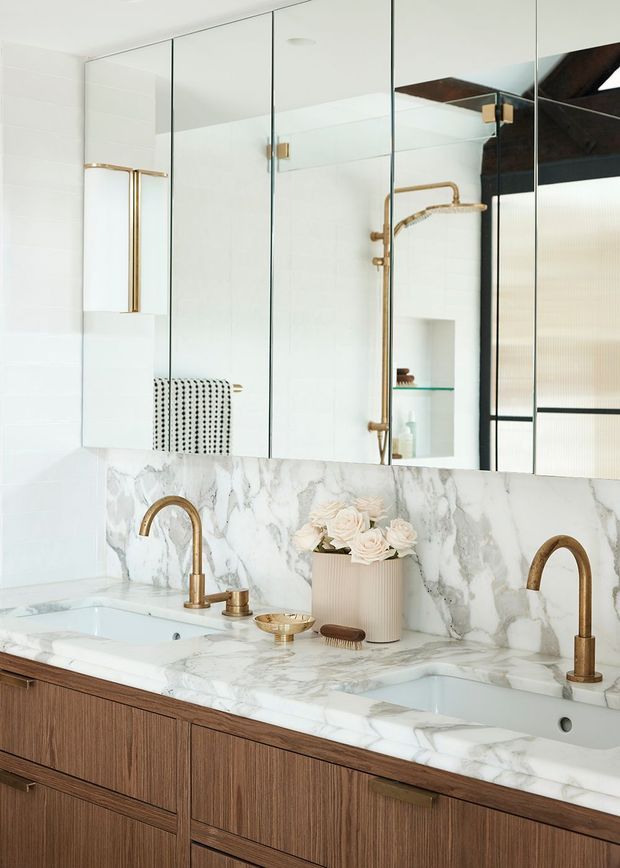 A refined double-vanity bathroom featuring a white marble countertop and backsplash with grey veining, warm wood-grain cabinetry, brushed brass faucets and shower hardware, and a full-width mirrored wall. A glass shower enclosure and recessed niche are visible in the reflection. The bright, polished scheme combines classic marble with contemporary custom joinery.