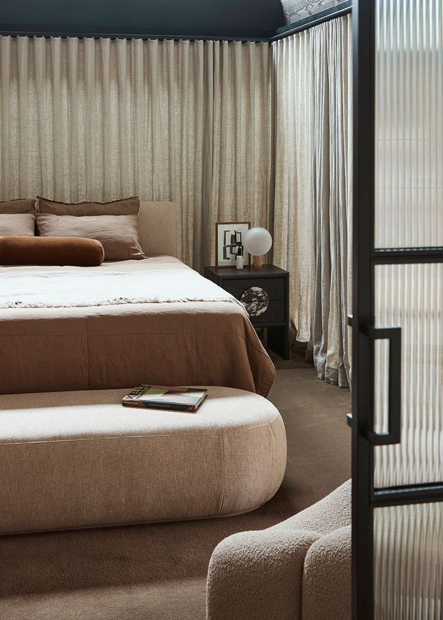 A warm, softly lit bedroom with floor-to-ceiling textured beige curtains, a low upholstered bed in taupe and brown tones, a curved upholstered bench at the foot, and a matching rounded lounge chair in the foreground. A dark bedside table with a spherical lamp and framed artwork adds contrast, while a black-framed fluted-glass partition or door partially screens the right side.
