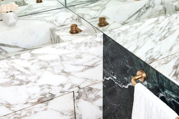 Close-up of a luxurious marble bathroom vanity or basin area, featuring heavily veined white and grey marble surfaces, a dark veined marble side panel, brass tapware, and a brass towel holder with a white towel. A mirror reflects the marble and fittings.