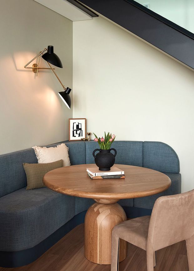 A compact, elegantly styled dining nook beneath a sloping ceiling. A curved blue upholstered banquette wraps around a rounded timber pedestal table, paired with a taupe upholstered chair. The space features warm timber flooring, cream walls, a sculptural black-and-brass wall lamp, cushions, framed artwork, and a small floral arrangement.