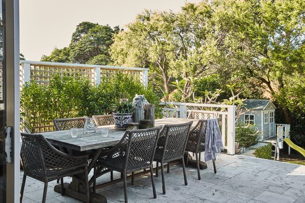 A sunlit outdoor dining terrace with a large rectangular dining table and woven dark chairs, bordered by white lattice privacy screens and lush mature planting. Pale stone paving extends toward a small light-coloured garden structure and play area in the background. The setting feels relaxed, leafy, and residential.