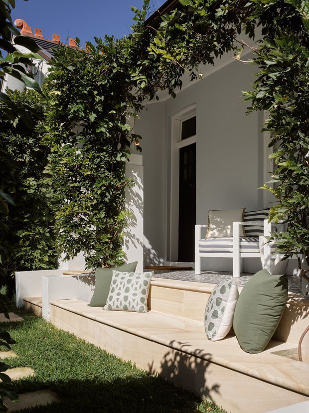 Sunlit landscaped courtyard terrace adjoining a white rendered building, framed by dense climbing greenery and garden planting. Broad pale stone steps form built-in bench seating with neutral and muted green cushions, while a white outdoor armchair sits on the upper landing beside a dark glazed doorway. A tiled roof edge is partially visible above the foliage.