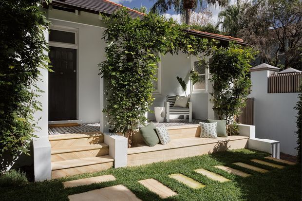 A leafy residential courtyard and entry terrace with a rendered white exterior, dark timber front door, tiled pitched roof, timber decking, built-in pale stone or concrete seating, and lush climbing greenery. Stepping stones lead through the lawn to the raised porch, which is furnished with cushions and a small outdoor chair.