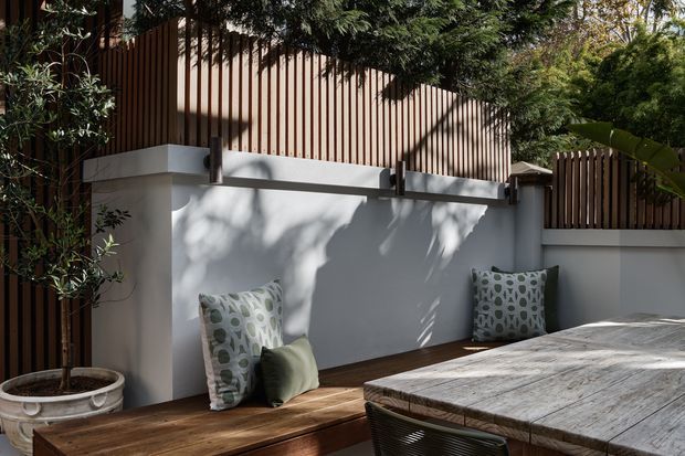 A shaded outdoor terrace with a built-in timber bench running along a pale rendered privacy wall. Vertical timber screening and mature planting create a secluded courtyard-like setting, while patterned cushions and a large weathered timber outdoor table add relaxed character.