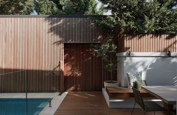 A refined outdoor courtyard entertaining area with a narrow swimming pool, timber decking, a vertical timber privacy screen and gate, and a built-in white-rendered bench with cushions beside a dining table. Mature planting provides dappled shade, while the warm timber and neutral finishes create a private, contemporary setting.