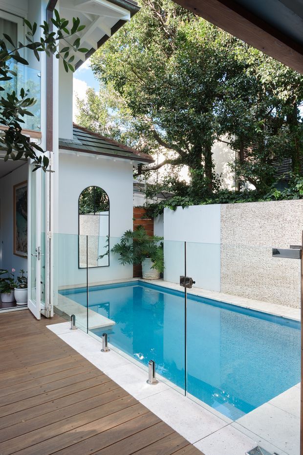 A compact outdoor swimming pool viewed from a timber deck beside a contemporary home. The pool is enclosed by frameless glass safety fencing and bordered by pale stone or concrete coping. White rendered walls, lush mature greenery, potted plants, and a partly open glazed folding door create a private, leafy courtyard atmosphere. A small outbuilding with a pitched roof and arched mirror is visible beyond the pool.