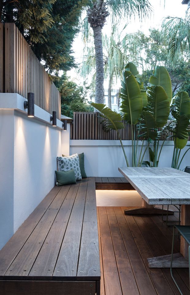 A compact landscaped outdoor courtyard or terrace with dark timber decking, built-in timber bench seating, white rendered walls, vertical timber privacy screens, black cylindrical wall lights, and lush tropical planting. A weathered timber dining table occupies the right side, while warm concealed lighting illuminates a recessed section beneath the bench.