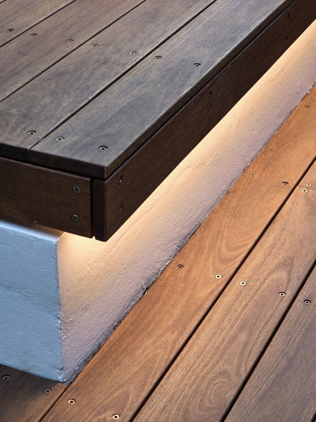 Close-up detail of dark stained timber decking and a projecting timber-clad platform or bench, with visible countersunk fixings and warm concealed LED lighting along the edge. A pale rendered wall and blue-grey painted structural element are partially visible.