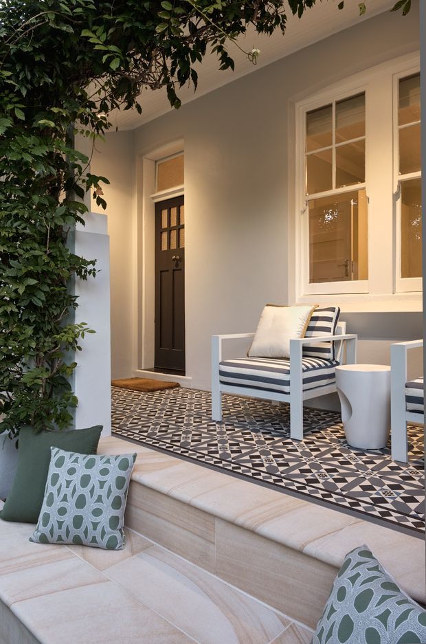 A refined outdoor porch or entry terrace with a patterned geometric tile floor, pale grey walls, white-framed windows, and a dark paneled entry door. White-framed striped lounge chairs and a small sculptural side table sit beside a built-in pale upholstered seating area with green patterned cushions. Dense leafy planting frames the space, creating a sheltered courtyard-like atmosphere.