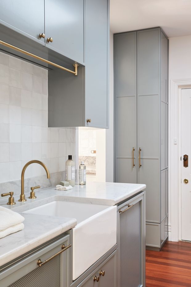 A compact, traditionally styled kitchen with a pale grey-blue colour scheme. The room features shaker-style cabinetry, tall pantry storage, brass knobs and pulls, a brass bridge-style faucet, white farmhouse apron-front sink, white marble-look benchtops, and a light square-tile splashback. A white paneled door and warm timber flooring are visible.