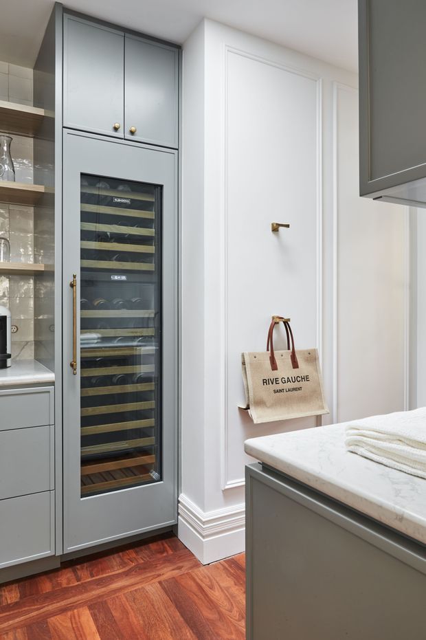 A refined kitchen interior featuring a tall grey built-in wine refrigerator with glass door and brass hardware, open shelving with glassware, grey cabinetry, white panelled walls with decorative trim, and a light marble-look island benchtop. Rich medium-toned timber flooring adds warmth and contrast.