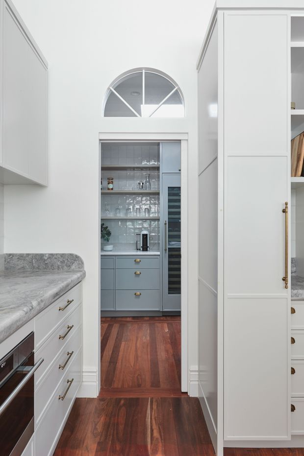 A bright, refined kitchen with white paneled cabinetry, grey stone benchtops, brass hardware, and dark reddish timber flooring. The view looks through a doorway into a compact butler’s pantry or scullery with blue-grey cabinetry, open glassware shelving, a tiled backsplash, and a wine fridge. A high arched internal window adds architectural character above the opening.
