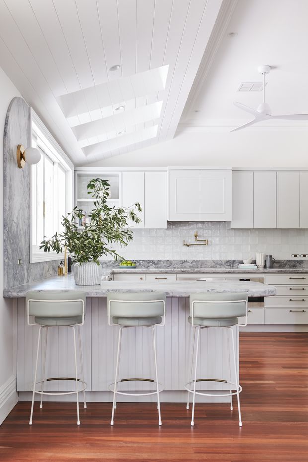 Bright, refined kitchen with a large island and three bar stools. The space features white shaker-style cabinetry, a grey-white marble benchtop and matching splashback, vertical panelled island joinery, warm timber flooring, extensive glazing, and a white tongue-and-groove ceiling with skylight-like panels. A contemporary ceiling fan, brass wall light, and indoor plant add detail.