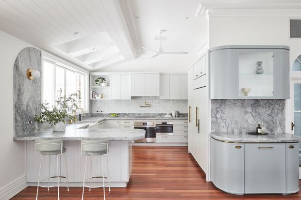 Bright, refined kitchen with white shaker-style cabinetry, a large marble-look island, matching stone splashback and feature wall, warm timber flooring, glass-front display cabinets, and two bar stools. The room includes abundant glazing, a partially vaulted panelled ceiling with skylights, and understated brass hardware.
