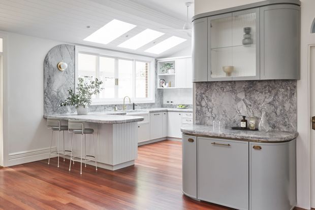 Bright, refined kitchen with a central island and two stools, pale grey cabinetry, extensive veined marble surfaces, integrated open shelving, and a curved built-in display and storage unit. Large windows and skylights provide abundant natural light, while warm timber flooring adds contrast.