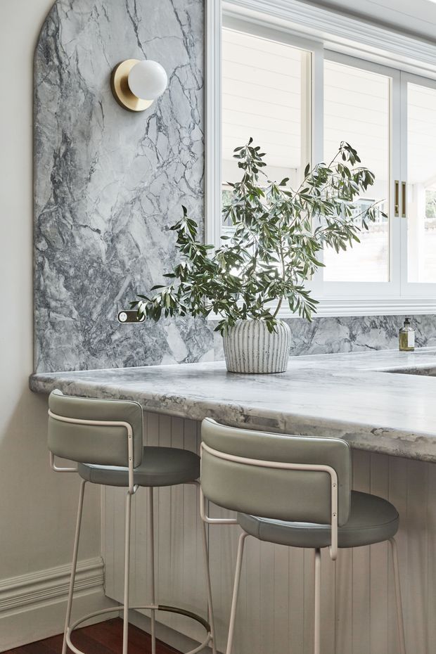 A refined, light-filled kitchen or entertaining space featuring a marble-clad wall and thick marble benchtop, white framed windows, vertical panelled cabinetry, and two upholstered counter stools. A sculptural wall sconce and potted leafy branch add understated decorative detail.