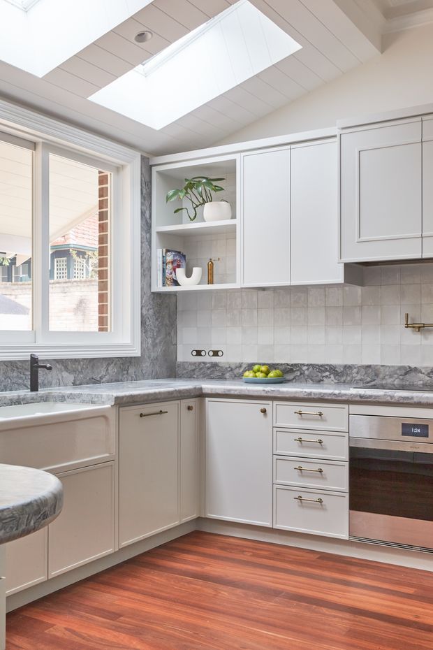 Bright, compact kitchen with white shaker-style cabinetry, grey marble-look benchtops and backsplash, a farmhouse sink, integrated oven, brass hardware and a large skylight. Warm timber flooring and a window with exterior views add contrast and natural light.