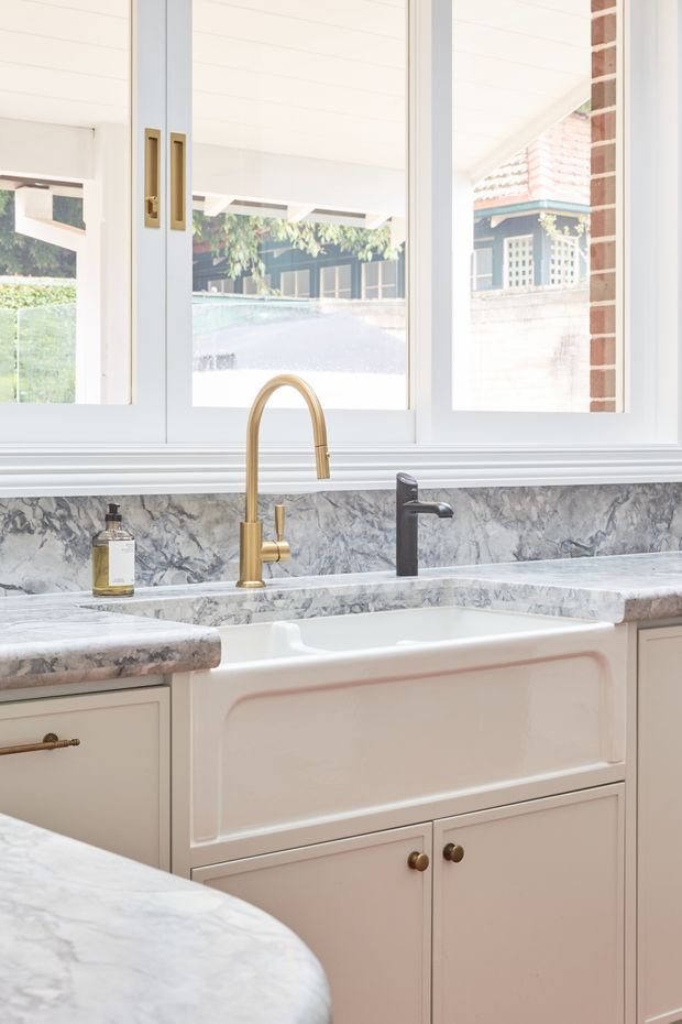 Bright traditional-style kitchen detail featuring a large white farmhouse apron-front sink, marble-look stone benchtops and splashback, pale cabinetry with brass hardware, and two contrasting sink mixers. White-framed windows overlook a covered outdoor area, with exposed brick visible at the side.