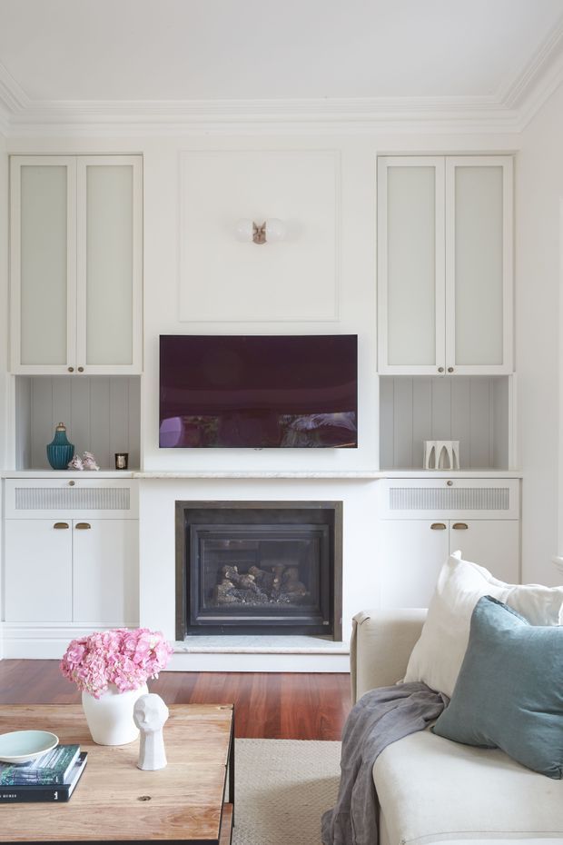 A bright, refined living room centred on a built-in white entertainment and fireplace wall. The room features symmetrical glass-front cabinetry, recessed panel detailing, a wall-mounted television, a dark fireplace surround, white painted joinery, timber flooring, and layered neutral seating with blue-green cushions. A timber coffee table and fresh floral arrangement add warmth to the otherwise light interior.