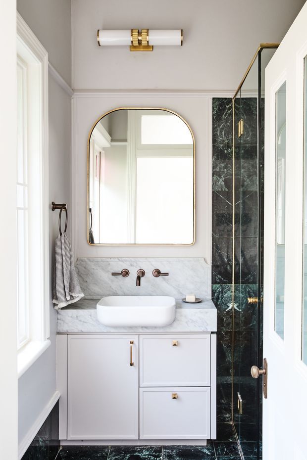 A compact, refined bathroom with a pale grey vanity, white vessel basin, marble countertop and splashback, dark veined marble wall and floor tiles, brass-framed arched mirror, and coordinated brass lighting and hardware. White panelled doors, trim, and a glass shower enclosure contribute to a polished classic look.
