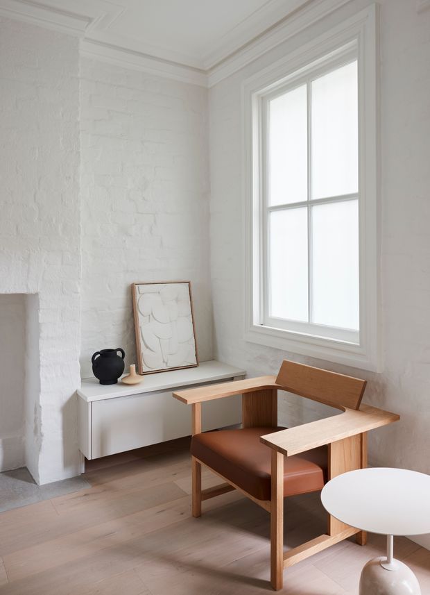 A bright, softly styled interior corner featuring white-painted exposed brick walls, detailed white cornices and trims, a large white-framed sash window, and pale timber flooring. A minimalist white console sits beneath the window with a framed artwork and black ceramic vessel, while a mid-century-inspired timber armchair with brown leather upholstery occupies the foreground beside a round white table. The space feels calm, refined, and restrained.