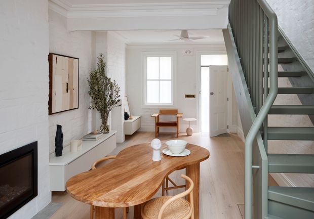 Bright open-plan interior combining a dining area and living space, with a sculptural timber dining table, pale timber flooring, white-painted brick walls, a compact fireplace, built-in console, and a green-painted staircase leading upward. The room has abundant natural light and a restrained contemporary coastal feel.