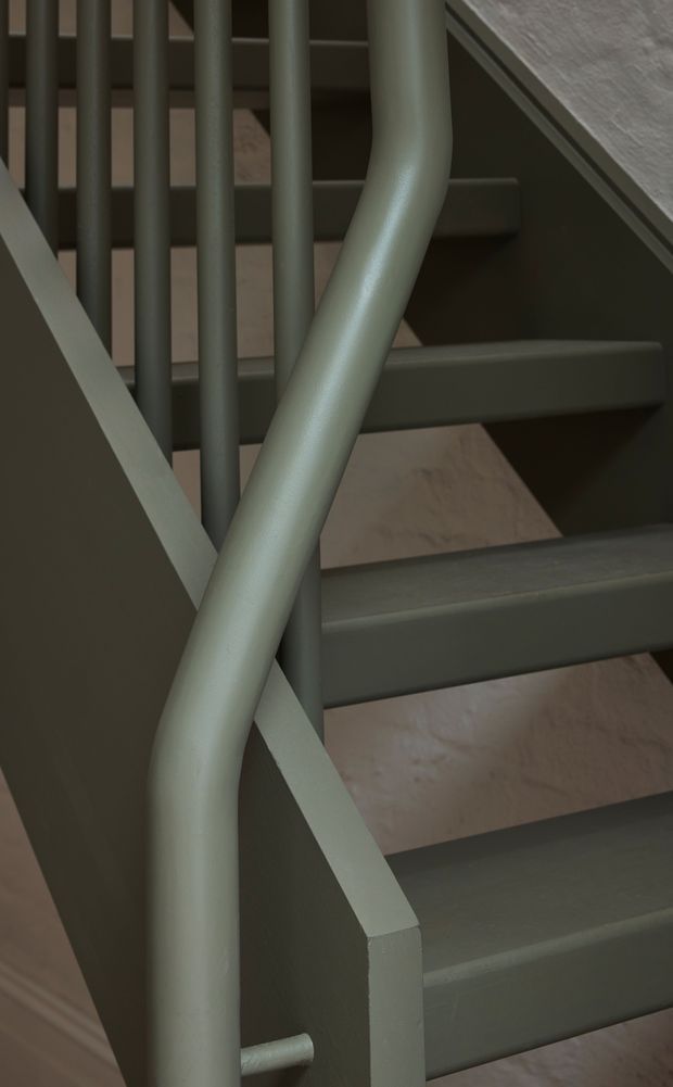 Close-up architectural detail of a staircase with a rounded, painted handrail, vertical balusters, solid side stringer, and dark painted stair treads. The muted green-grey stair structure contrasts with warm timber visible on the steps and surrounding surfaces.