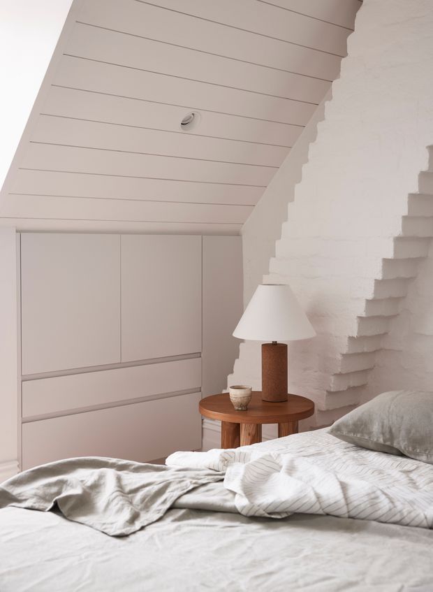 A softly lit bedroom with a low bed in the foreground, pale textured bedding, built-in light cabinetry, and a round timber bedside table with a ceramic lamp. The room features a white painted brick wall with a distinctive stepped, irregular edge and a white panelled sloping ceiling, creating a calm, warm minimalist interior.
