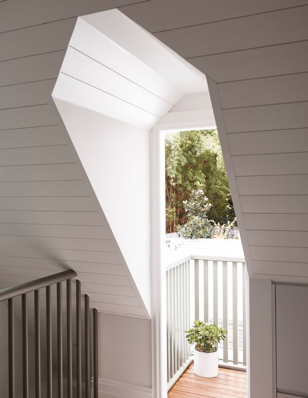 A bright interior stair landing with a dramatic angular opening leading to a small outdoor balcony. White horizontal wall and ceiling panelling surrounds the stairwell, with dark metal balustrades, a white-framed glazed opening, and timber decking outside. A potted plant sits on the balcony, with greenery visible beyond.