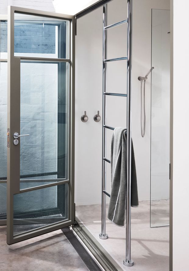 A compact contemporary bathroom with a frameless glass shower enclosure, large pivoting glass door, chrome ladder-style towel rail, handheld shower fitting, and pale concrete-look flooring. A full-height mirror and clean white walls create a bright, minimal space.