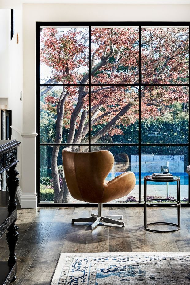 A refined interior seating area with a sculptural brown leather lounge chair positioned before expansive black-framed floor-to-ceiling glazing. The windows overlook a landscaped garden and swimming pool. Dark timber flooring, classical white wall and column mouldings, a black ornate cabinet or fireplace edge, a slim round side table, and a patterned area rug create a high-contrast, elegant setting.