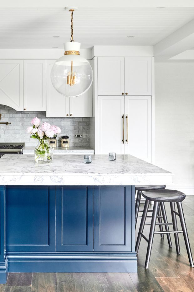 Bright, refined kitchen featuring a large white marble island with deep navy-blue paneled cabinetry, white shaker-style wall cabinets, a grey subway-tile splashback, dark timber flooring, and black stools. A prominent glass-and-brass pendant light hangs above the island, with understated floral styling on the benchtop.