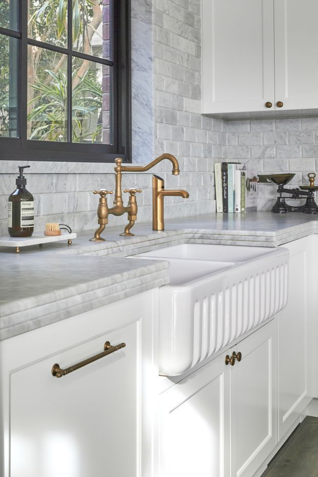 Close-up photograph of a refined traditional kitchen, featuring white shaker-style cabinetry, a large white ceramic farmhouse sink, pale grey veined stone benchtops, grey brick-pattern tiled splashback, dark framed window, and aged brass tapware and hardware. Natural light enters through the window, with greenery visible outside.