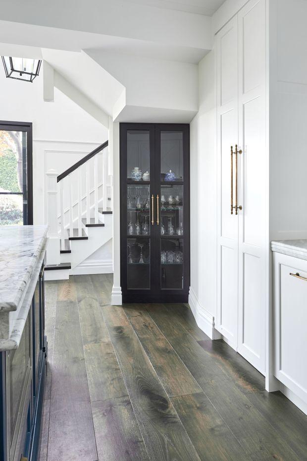 Bright, refined kitchen interior with white shaker-style built-in cabinetry, marble countertops, a dark glass-front display cabinet, and dark timber flooring. A staircase with white balustrades and dark handrail is visible beyond the kitchen, along with black-framed glazing and brass cabinet hardware.