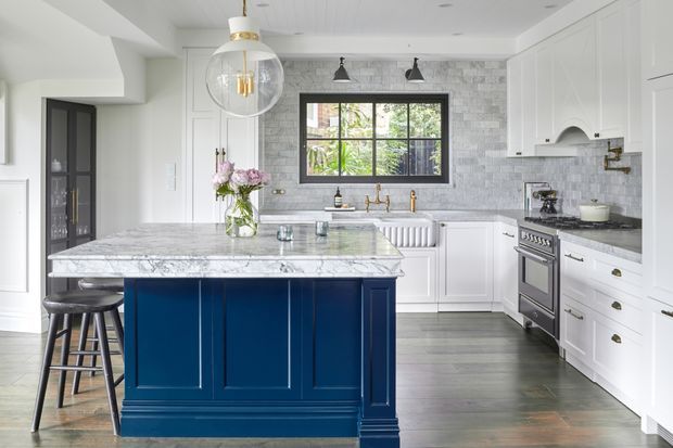 Bright, refined kitchen with a large navy-blue island topped in veined marble, white shaker-style cabinetry, a grey marble-tile splashback, dark timber-look flooring, brass hardware and fittings, and black-framed window and doors. Pendant and track lighting illuminate the island and cooking area.