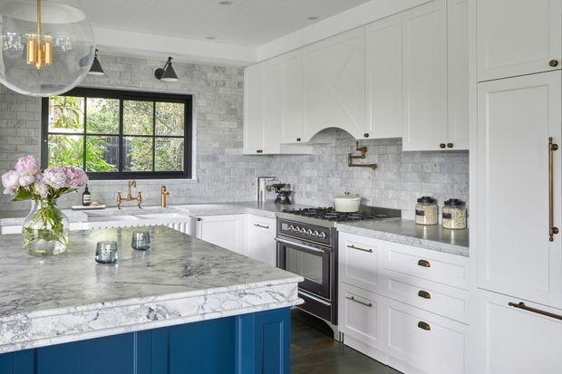 Bright, refined kitchen with white shaker-style cabinetry, a deep blue island, marble countertops, a pale stone-tile backsplash, and a stainless steel range. Black-framed windows, brass hardware and fixtures, pendant lighting, and floral styling add contrast and warmth.