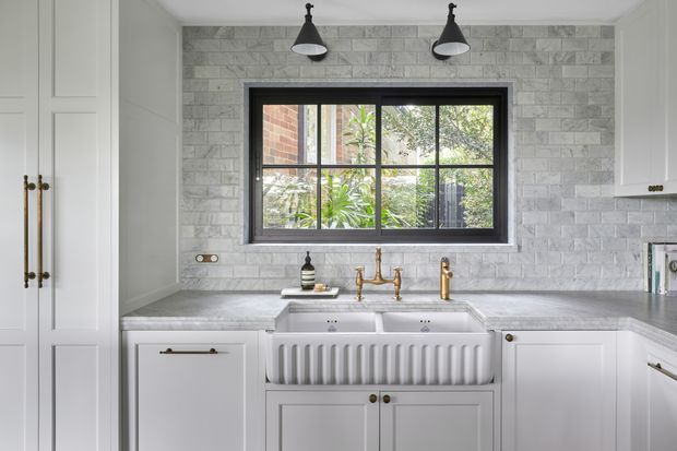 A bright, refined traditional kitchen with white Shaker-style cabinetry, a large apron-front farmhouse sink, grey stone benchtops, and a textured light-grey stone-tile splashback. A black-framed multipane window overlooks greenery, while two black pendant lights and brass tapware provide contrast.