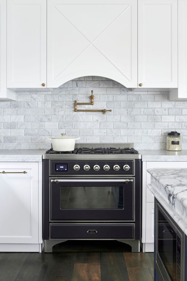 A bright, compact kitchen centered on a dark freestanding range cooker. White paneled cabinetry with small brass knobs surrounds a curved decorative rangehood canopy, while a light grey-white subway tile splashback and pale stone benchtops create a classic, refined look. A marble-look island or peninsula is visible at right, with dark timber flooring below and brass pot-filler plumbing above the stove.