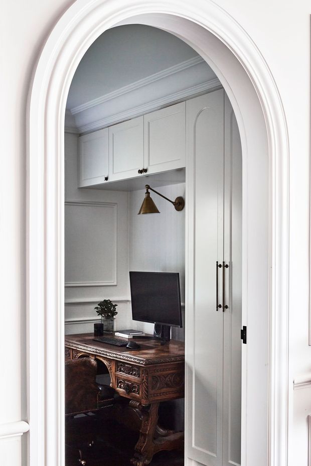 A compact home office viewed through an ornate white arched doorway. The room features built-in pale grey cabinetry with paneled doors, a traditional carved dark wood desk, leather chair, computer monitor, bronze wall lamp, decorative wall moulding, and crown cornice. The space has a refined traditional character with a strong contrast between light cabinetry and dark timber furniture.