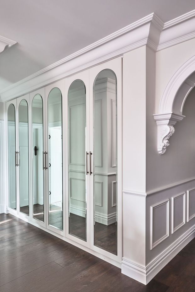 Elegant traditional interior hallway featuring a wall of tall arched mirrored wardrobe or storage doors with slim dark handles. The space has white decorative crown moulding, ornate architrave detailing, carved corbels, and raised wall panelling over dark timber flooring. The mirrors reflect additional panelled interior surfaces and doors.