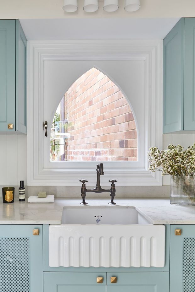 Bright, highly detailed kitchen sink vignette featuring a white ceramic farmhouse apron-front sink beneath an unusual pointed-arch window. Pale blue-green shaker cabinetry with brass knobs frames a light stone benchtop, while a dark aged-metal bridge tap and white decorative wall mouldings add traditional character. A brick wall is visible through the window, with flowers and small countertop accessories providing styling.