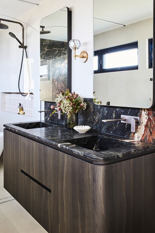 A refined contemporary bathroom vanity with a double dark stone basin, dark timber-grain cabinetry, and a matching dramatic stone splashback. Two rounded rectangular mirrors sit above the vanity, complemented by brass wall sconces and dark-framed horizontal windows. A walk-in shower with a dark metal shower set is visible to the left. The space combines dark timber, richly veined stone, pale wall tiles, and warm metallic accents.
