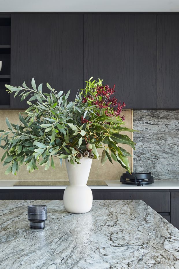 Close-up view of a refined contemporary kitchen, dominated by a large veined grey-and-white stone island benchtop in the foreground. Dark vertically grained cabinetry spans the rear wall, with a warm neutral stone or tiled splashback and matching stone work surface at the cooktop. A sculptural white vase with leafy branches and red berries provides a natural focal point, alongside a dark cooktop and ribbed glass tumbler.
