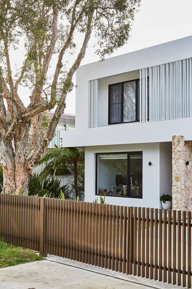 Street-facing view of a contemporary two-storey residence with a clean white rendered facade, dark-framed windows, vertical privacy screens, a timber-toned slatted front fence, and a stone-clad entrance wall. Mature trees and tropical planting frame the property.
