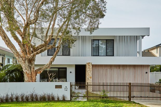 Front exterior view of a contemporary two-storey residence with a flat roof, crisp white rendered volumes, vertically ribbed metal cladding across the upper level, and a stone-clad entry column. The property has a low white boundary wall, timber-toned metal fencing and gate, large dark-framed windows, and established landscaping including a prominent street tree.