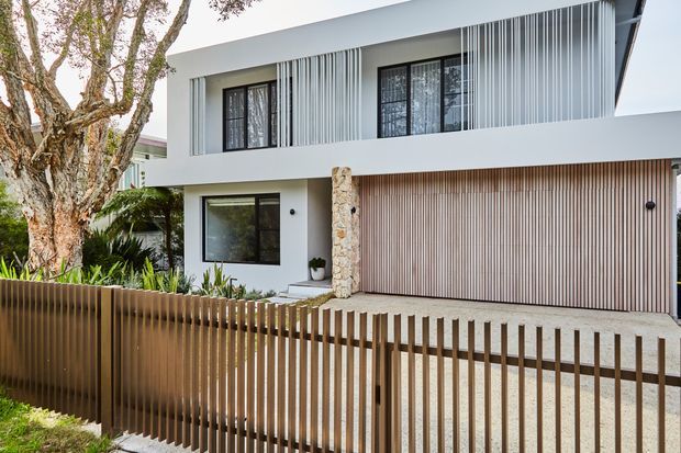 Street-facing view of a contemporary two-storey residence with a flat roof, crisp white rendered walls, dark window frames, vertical metal screening on the upper facade, and a warm timber-look slatted garage door. A stone-clad entry column, landscaped front garden, and vertical slat boundary fence complete the facade.