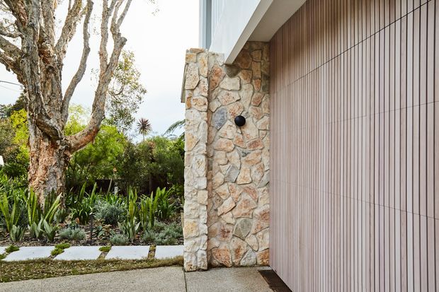 Exterior view of a contemporary home entrance, featuring a natural irregular-stone wall, a vertically grooved light-toned facade panel, and a covered soffit. A concrete path and lush landscaped garden with drought-tolerant planting lead toward the entry.
