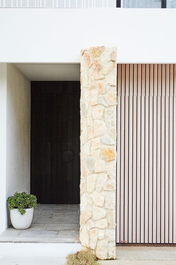Close-up exterior view of a contemporary residential entry, featuring a recessed dark timber door, pale grey tiled threshold, a prominent rough natural-stone column, white rendered wall surfaces, and a vertically slatted timber garage door. A potted plant sits beside the entrance.